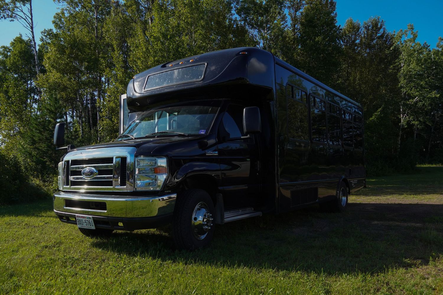 Black Ford party bus parked on green grass surrounded by tall trees on a sunny day.