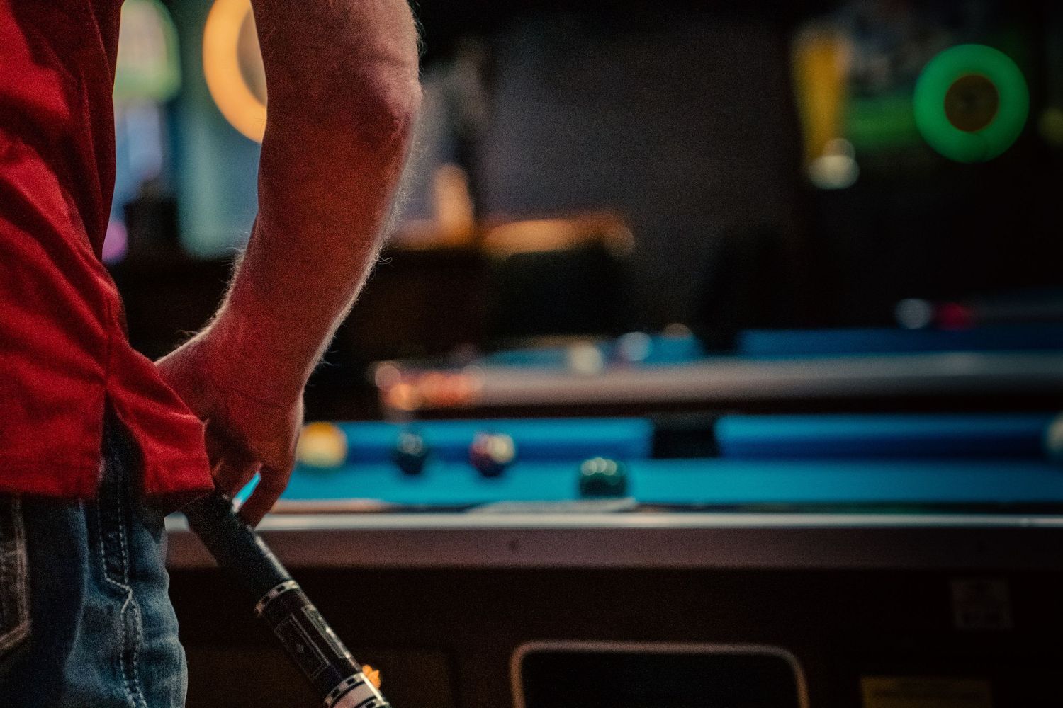 Man in red shirt holding a pool cue near a billiard table with balls in a dark bar setting.