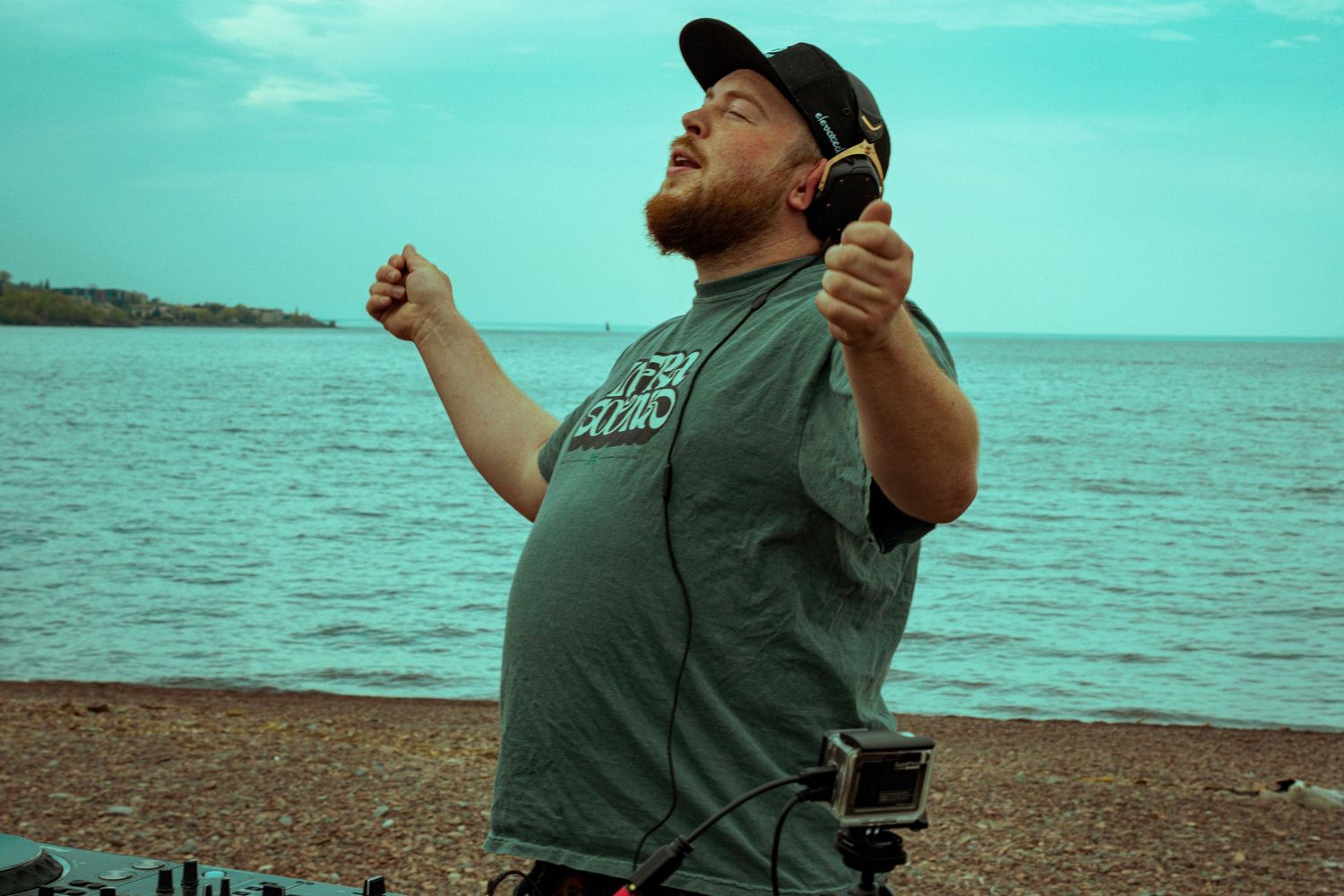 Man in gray t-shirt holding headphones poses joyfully on a rocky beach with calm turquoise water behind him.