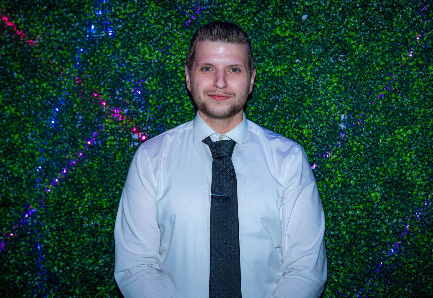Young man in white dress shirt and dark tie posing in front of a green ivy wall with twinkling lights.
