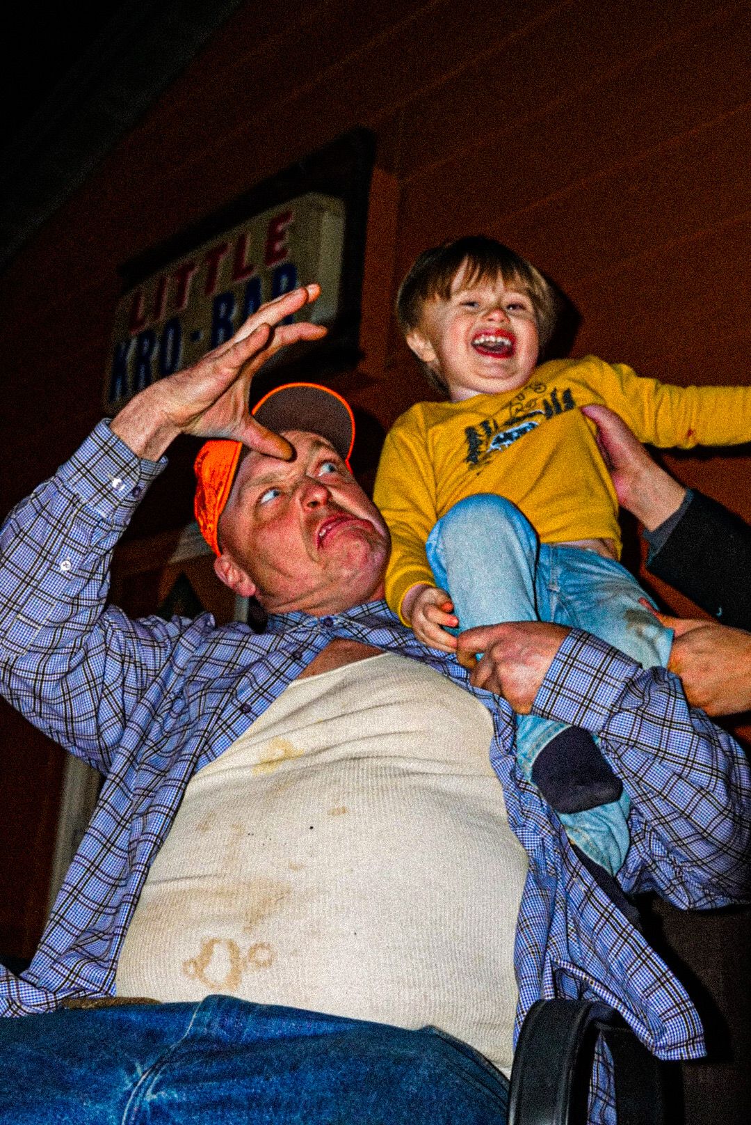 Man in plaid shirt and orange cap playfully holds laughing toddler in yellow shirt outdoors at night.