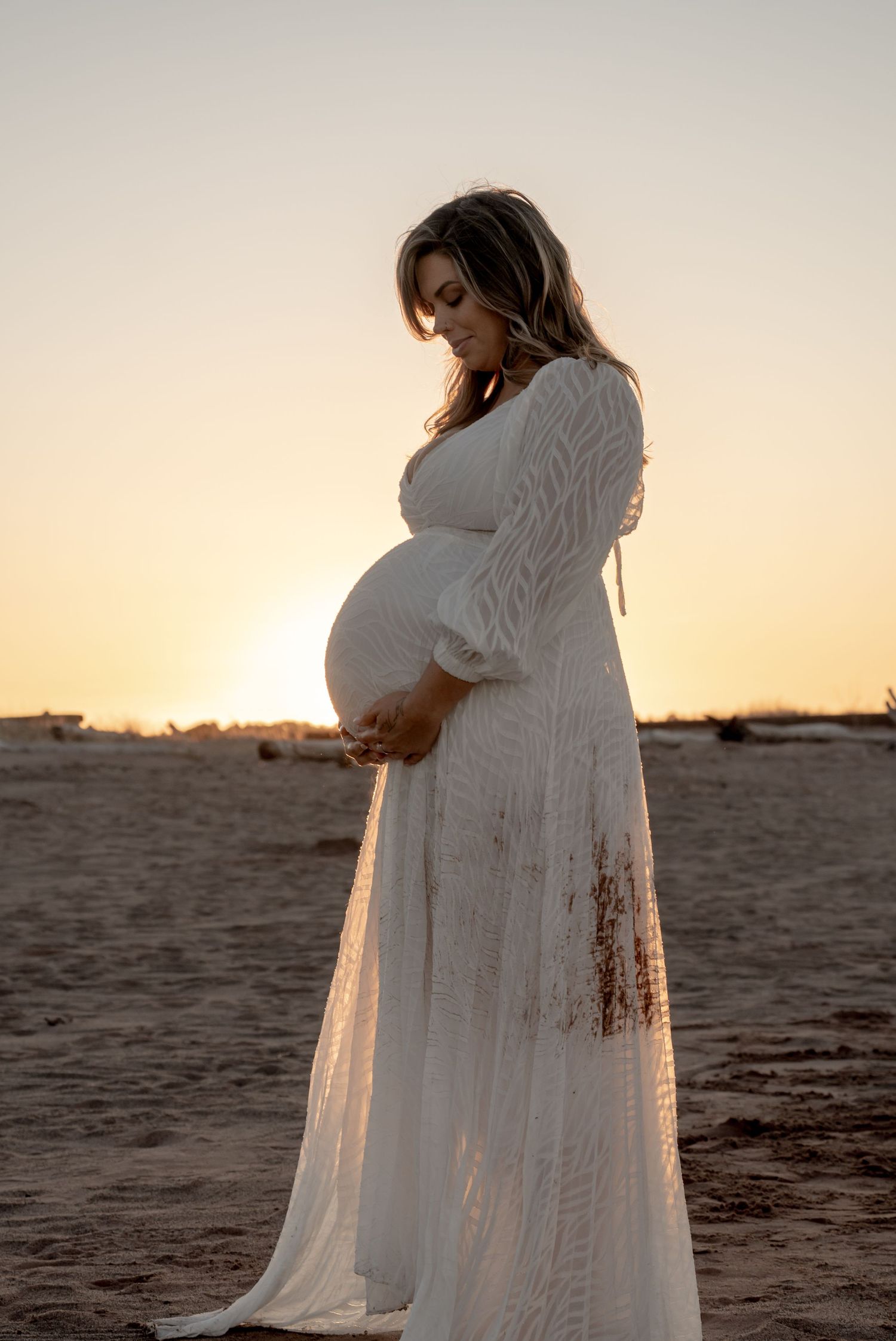 Pregnant woman in white lace gown cradling belly at sunset on sandy beach during golden hour maternity photoshoot.