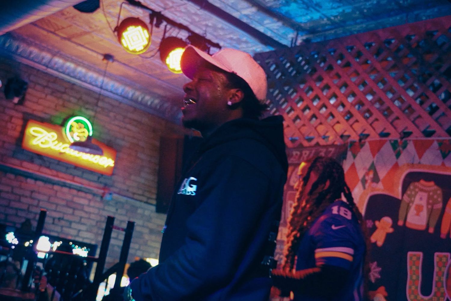 Performer in white hat and dark hoodie on stage at a dimly lit bar with Budweiser neon sign.