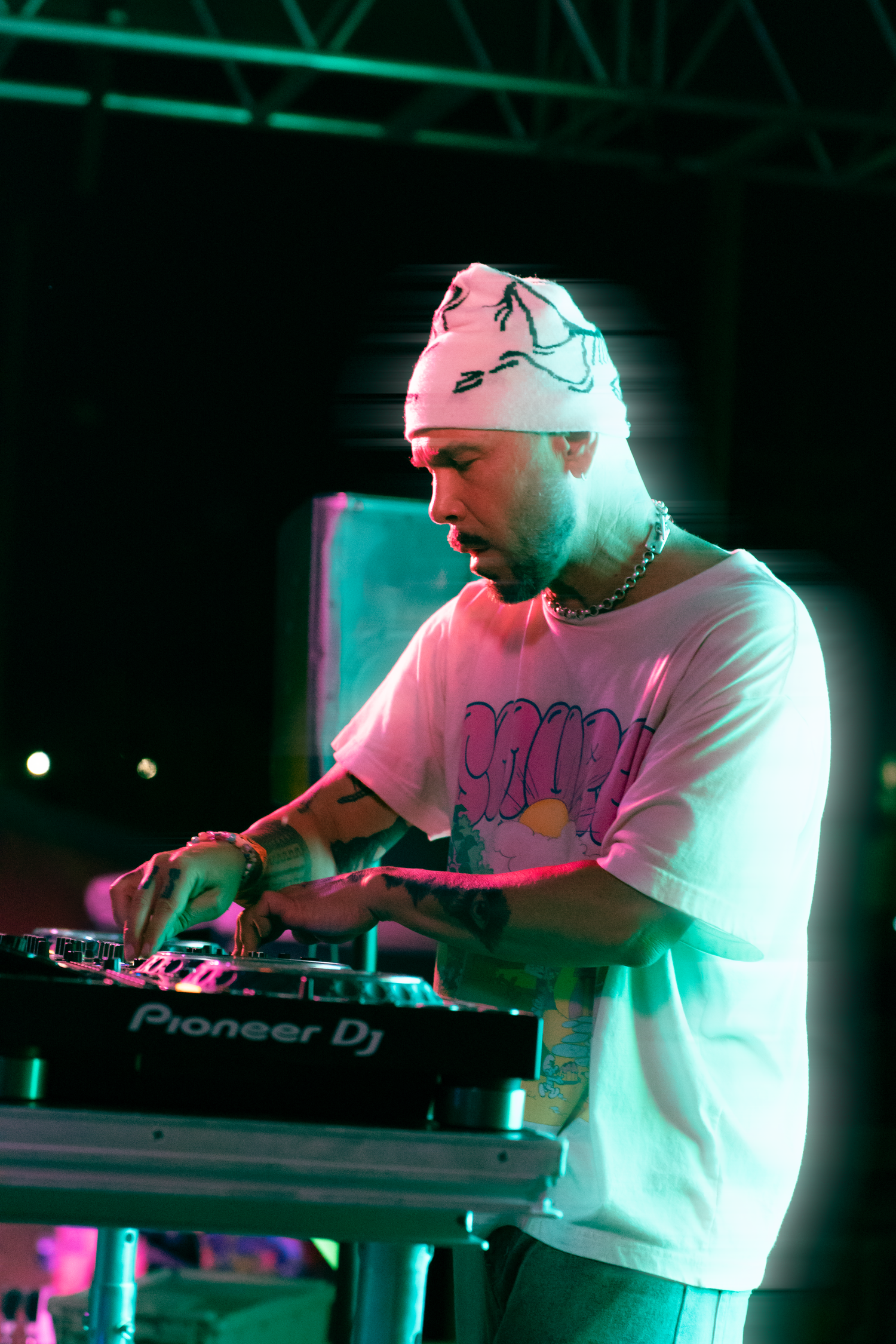 A DJ performs on stage using Pioneer DJ equipment under colorful pink and green stage lighting while wearing a white beanie.