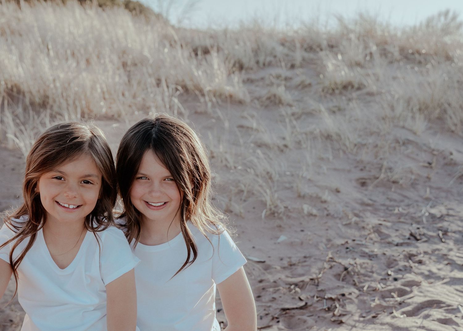 Two young girls with brown hair smile together on a sandy beach with golden dunes in the background.