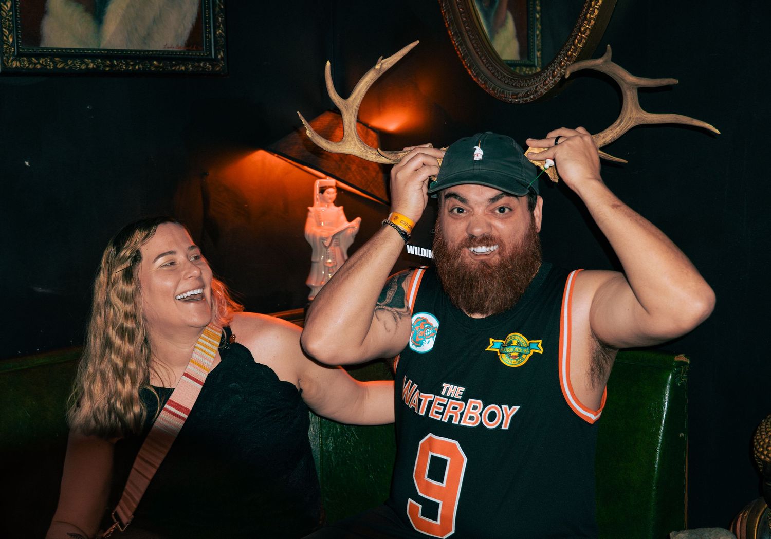 Man in Waterboy jersey and cap poses with antlers while laughing woman watches in dimly lit bar.