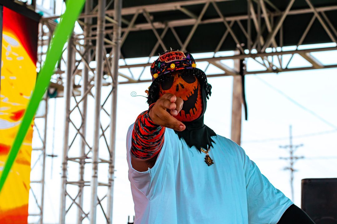 Performer in colorful tiger-print hat and dreadlocks points at crowd on outdoor festival stage.