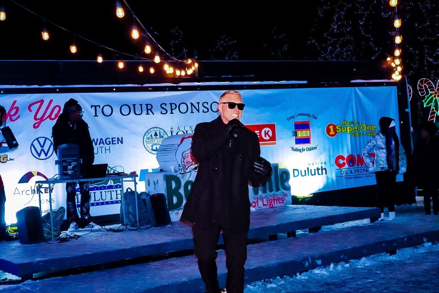 Man in black suit and sunglasses speaking at an outdoor winter event stage with sponsor banners at night.