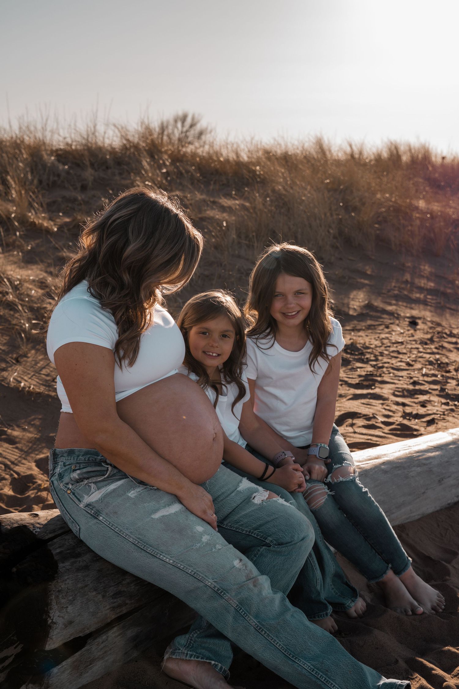 Pregnant woman sitting on driftwood at beach with two young girls in matching white tops and jeans on sunny day.