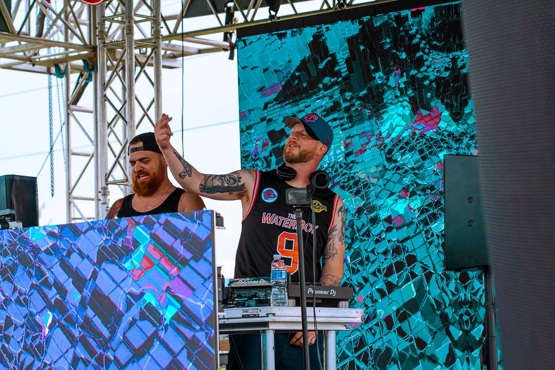 Two DJs performing on stage at an outdoor festival, raising hands with colorful LED screens behind them.