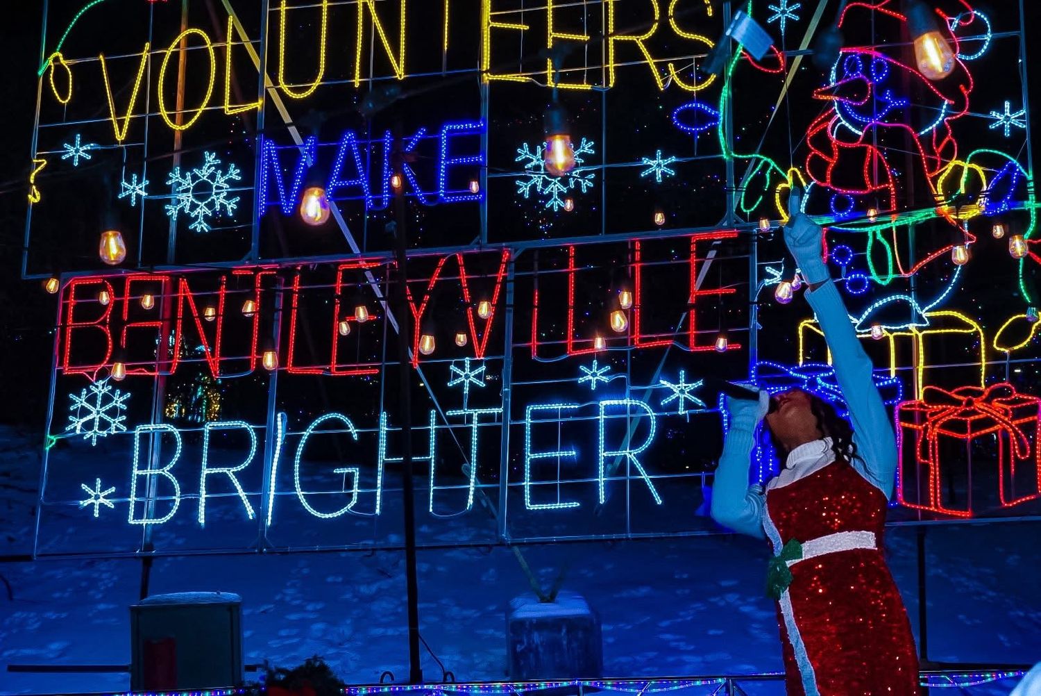Person in Santa suit poses before colorful Bentleyville holiday light display reading Volunteers Make Bentleyville Brighter.