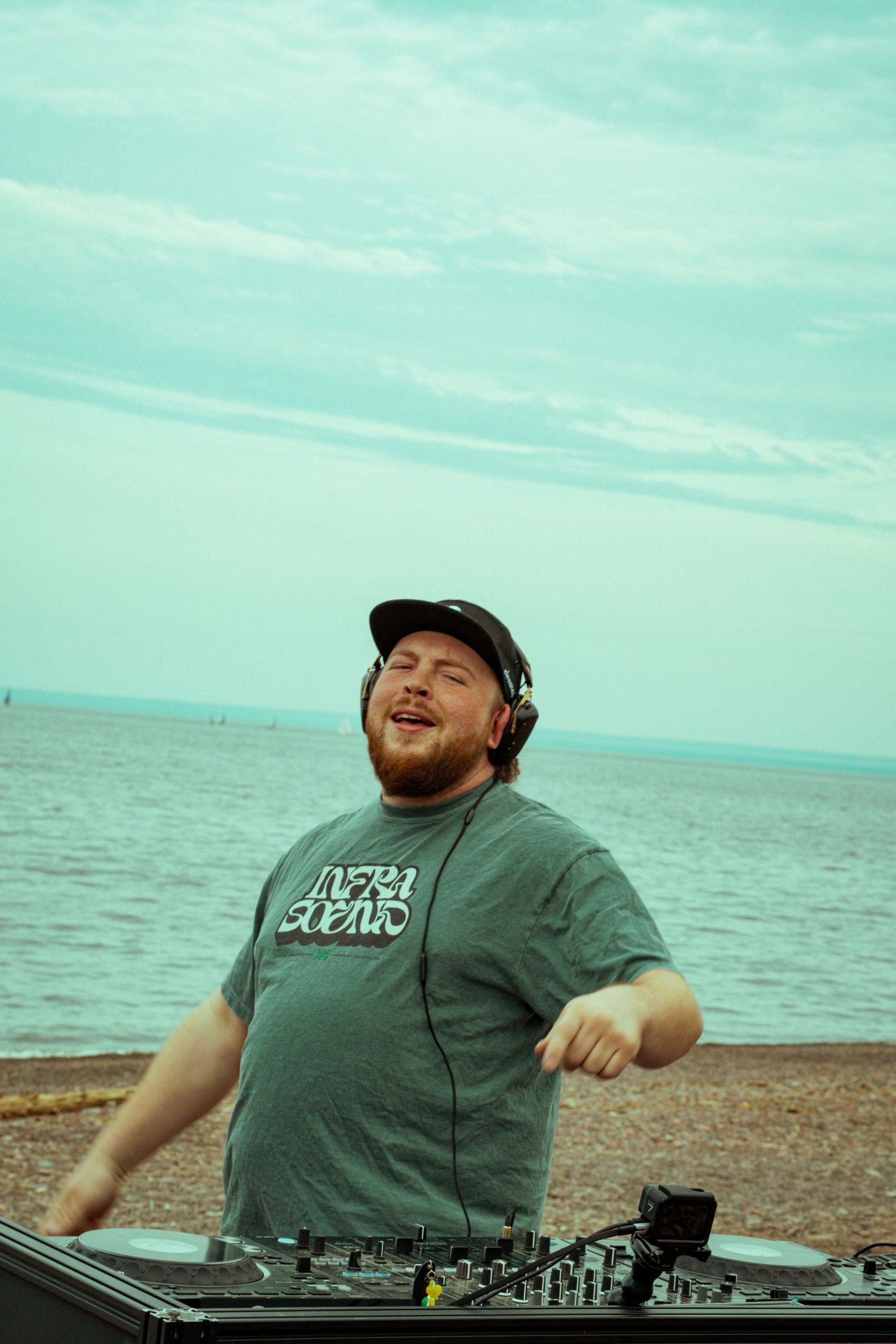 Bearded DJ wearing headphones and cap mixing music on turntables at a scenic beachside outdoor event.