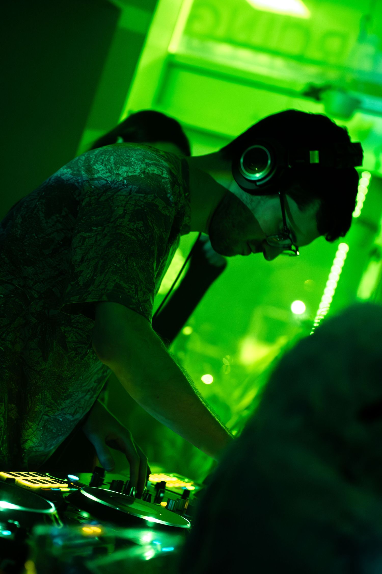 DJ with headphones and glasses leaning over turntables under green neon lights at a nightclub event.