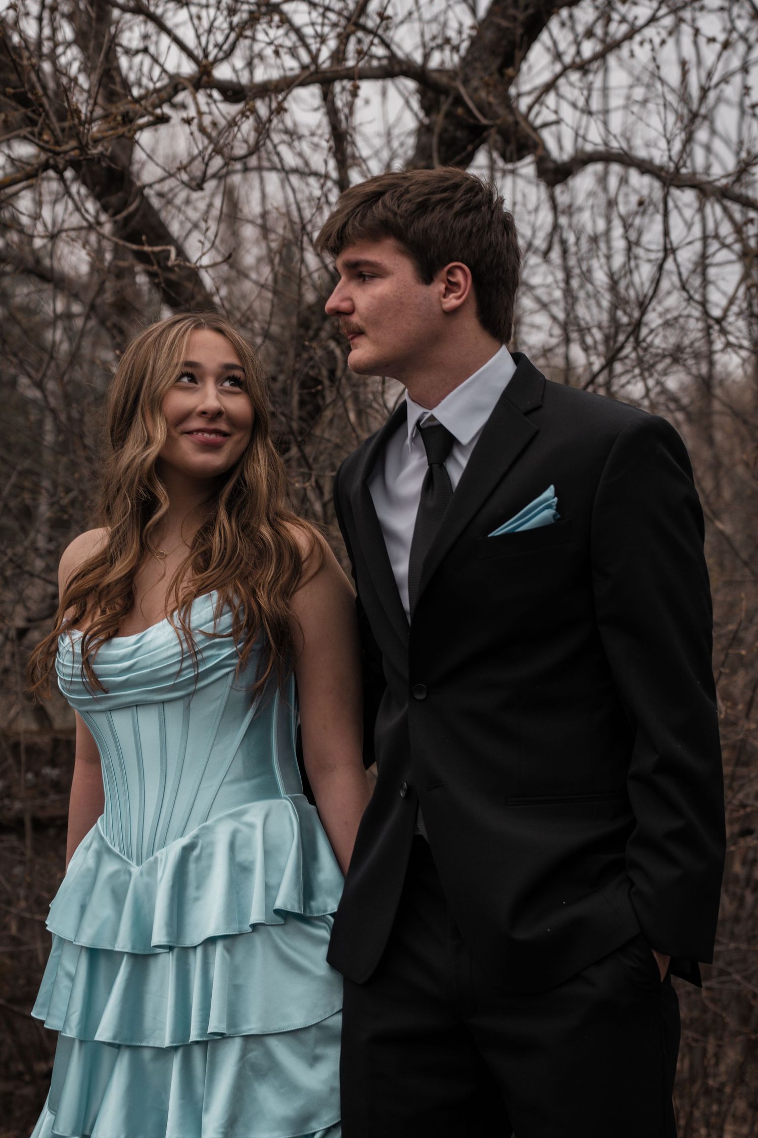 A young couple in matching light blue prom attire poses outdoors among bare winter trees.