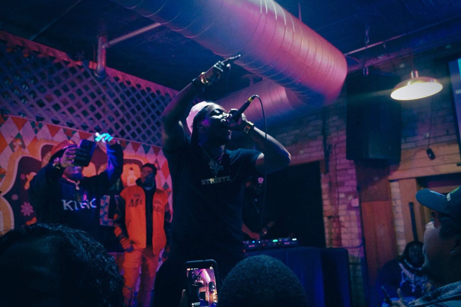 Rapper performing on stage with microphone raised in dimly lit venue with colorful lighting.