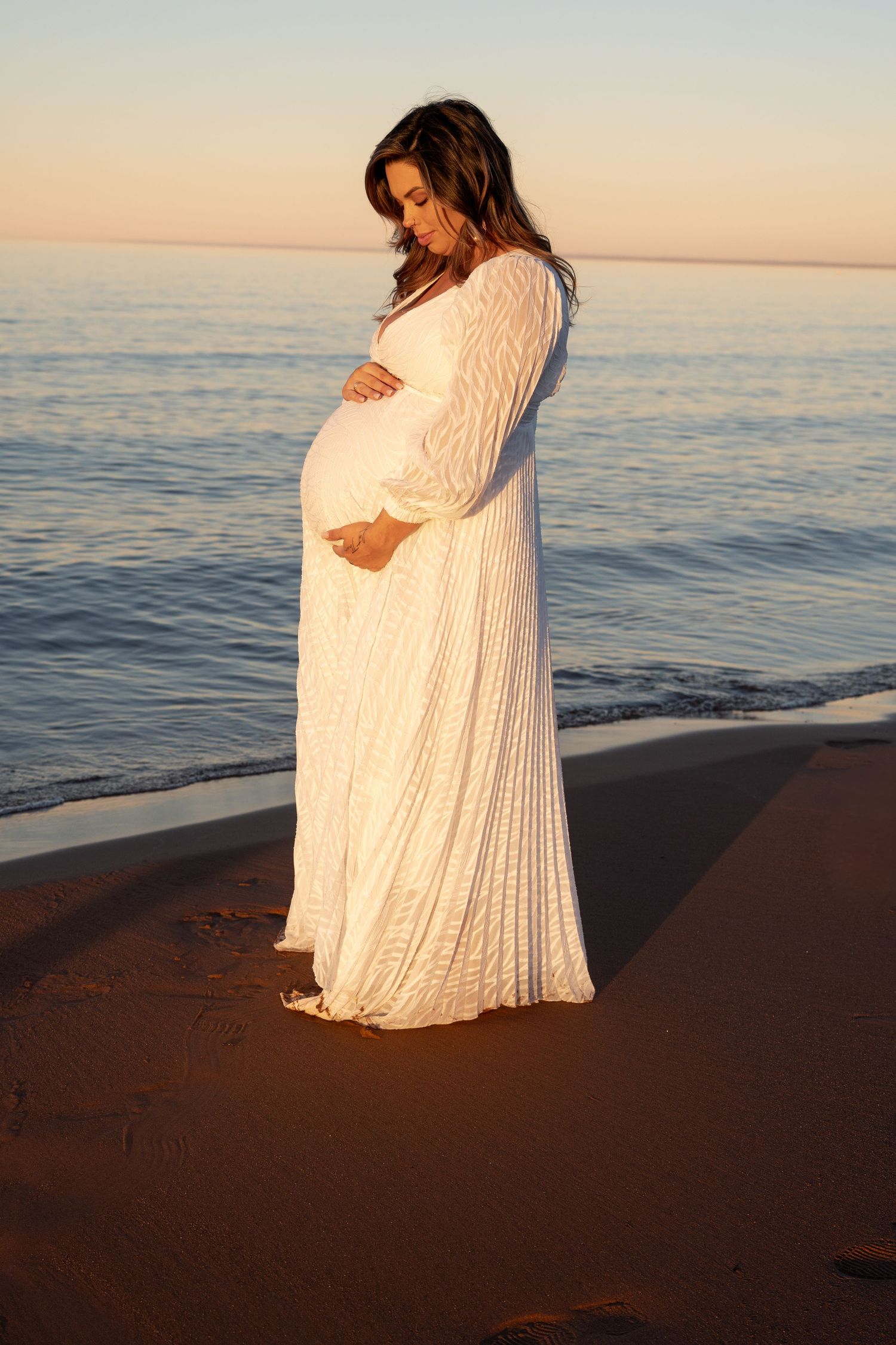 Pregnant woman in white lace gown cradling baby bump on sandy beach at golden sunset.