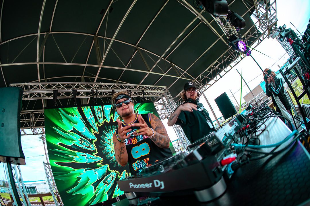 DJ performing on outdoor stage with Pioneer DJ equipment, green psychedelic backdrop, under metal truss canopy.