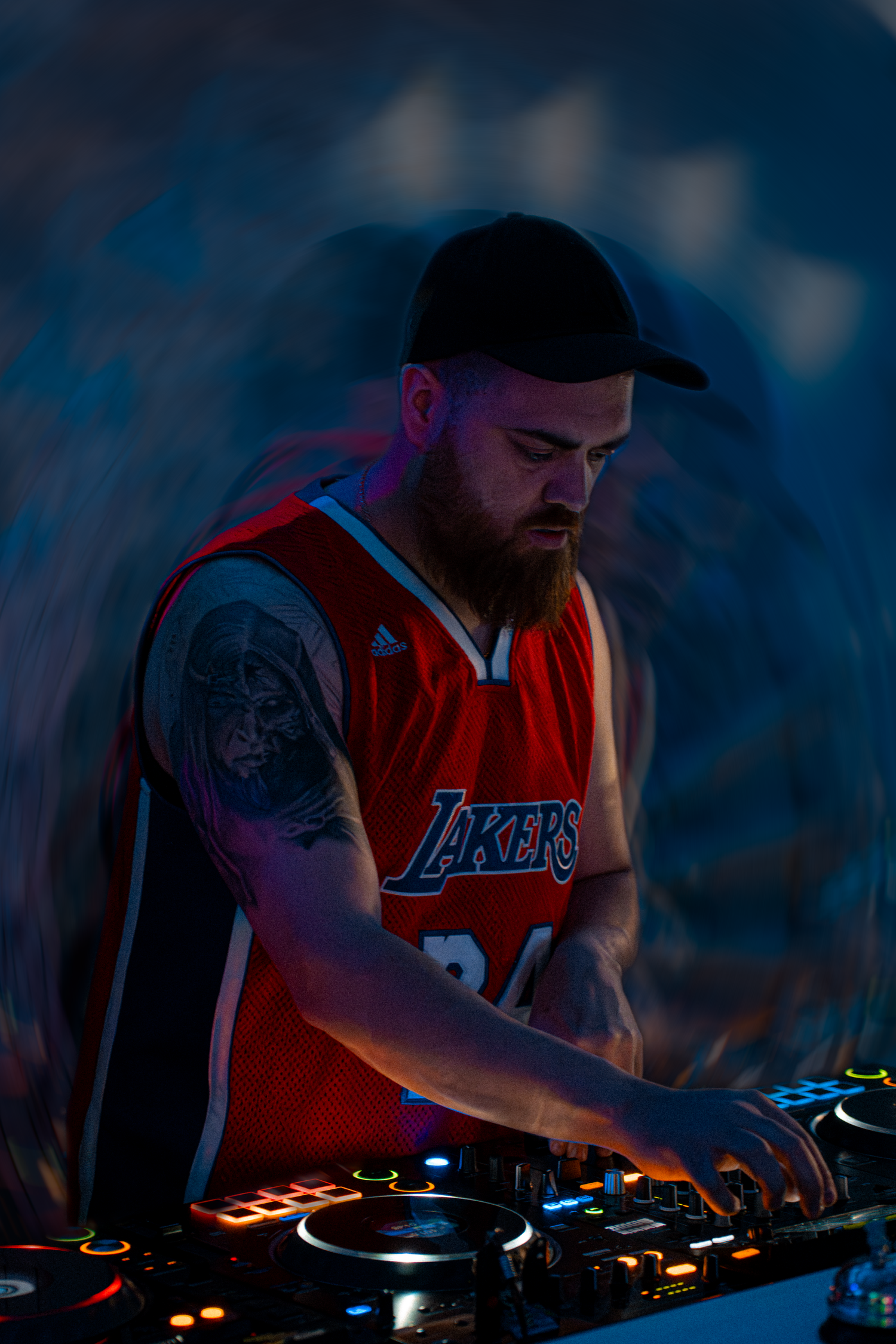 Bearded DJ in Lakers jersey mixing on turntables under blue and red stage lighting.