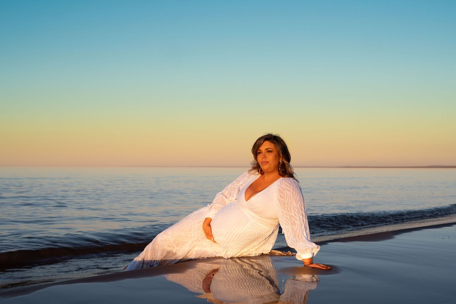 Pregnant woman in white dress sitting on beach shore at golden sunset with calm ocean waters reflecting warm light.