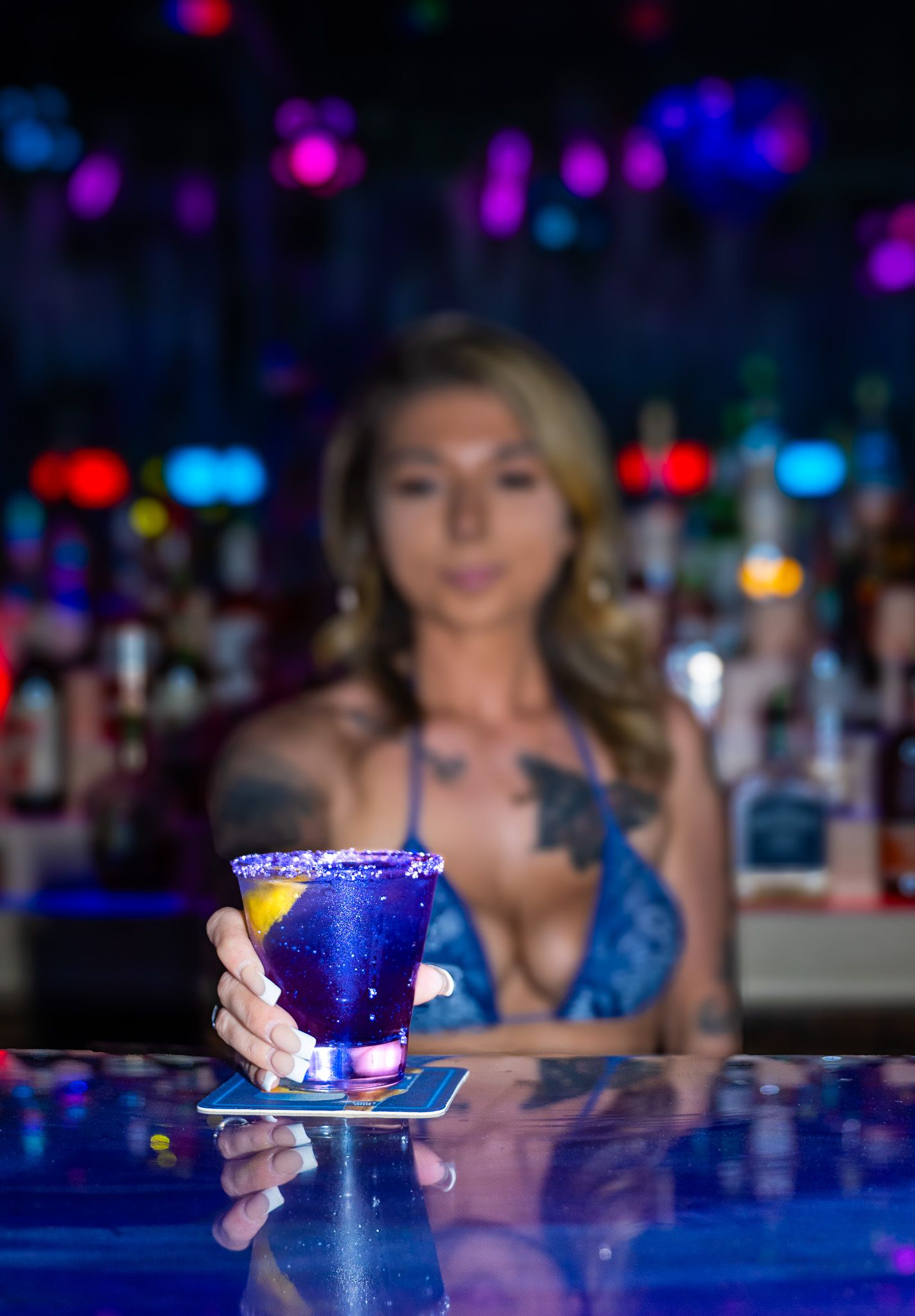 Tattooed female bartender holding a vibrant blue cocktail in a martini glass at a neon-lit bar.