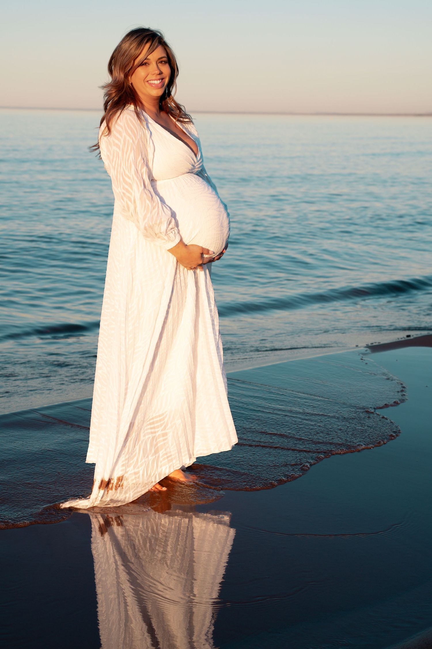 Pregnant woman in white dress standing barefoot on beach shore, cradling baby bump, with ocean reflection at sunset.