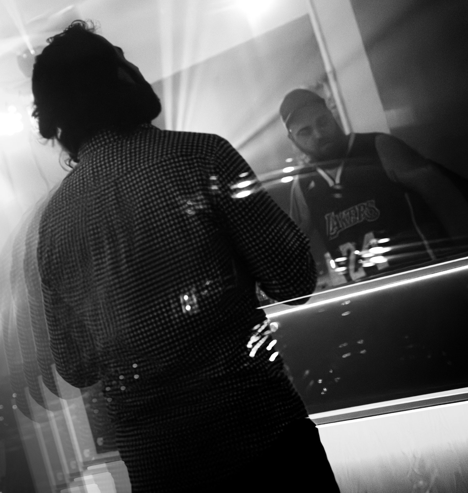 Black and white photo of person in sequined jacket viewed from behind, reflected in mirror with man in Lakers jersey.