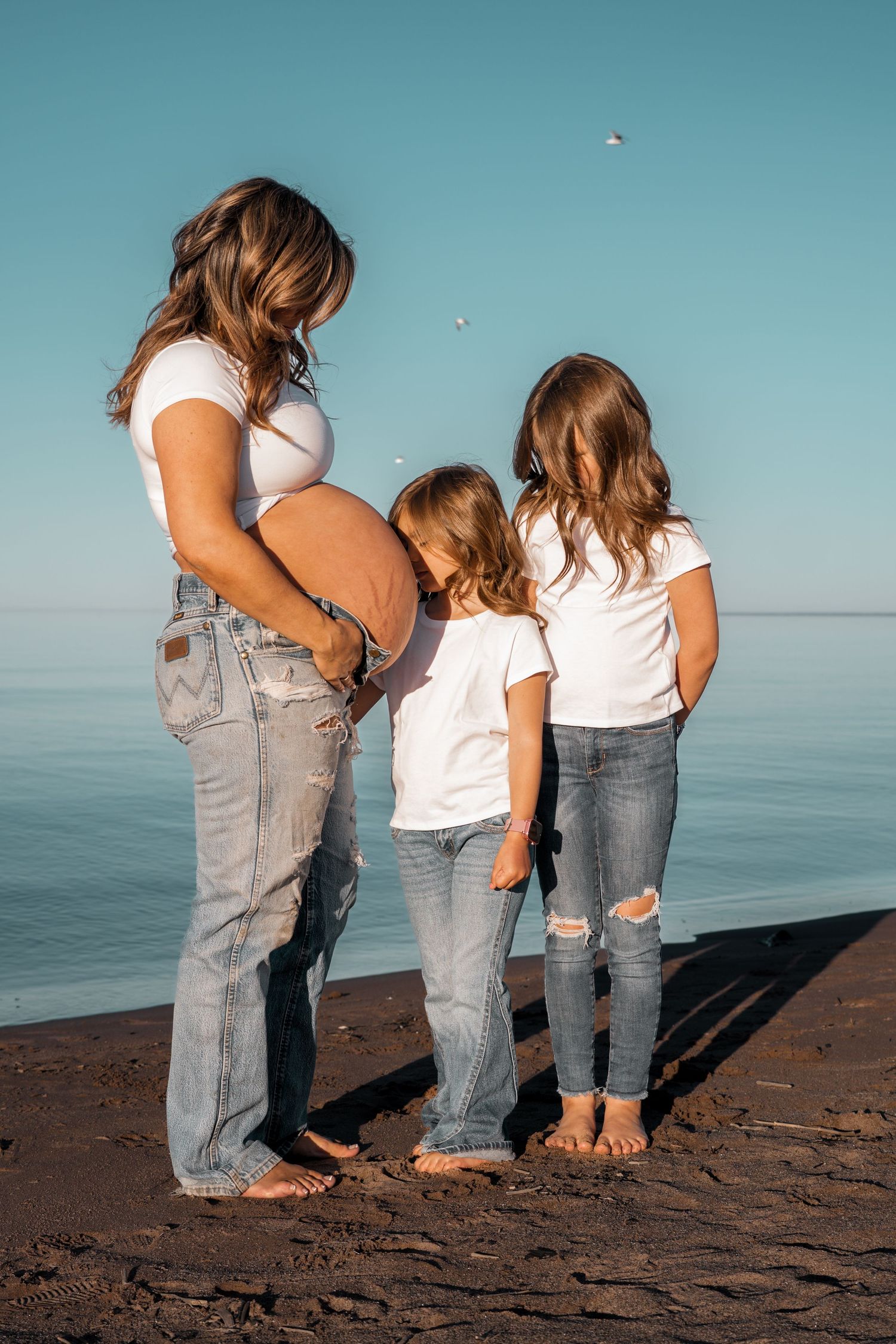 Pregnant woman in white top and jeans with two young daughters by the ocean on a sunny day.