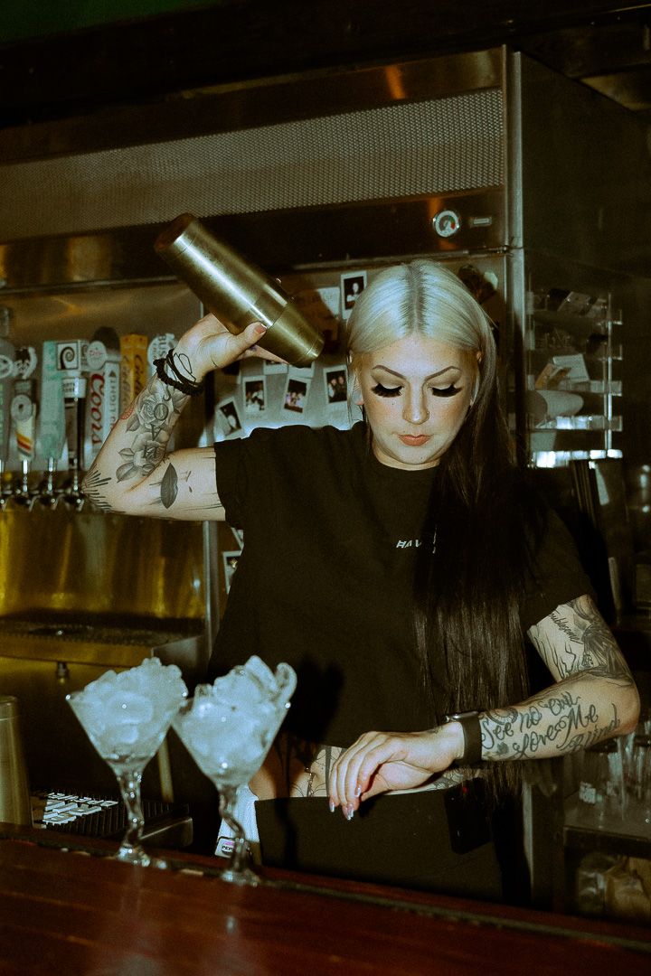 Tattooed female bartender in black shirt pouring cocktail shaker into martini glasses at a dimly lit bar.