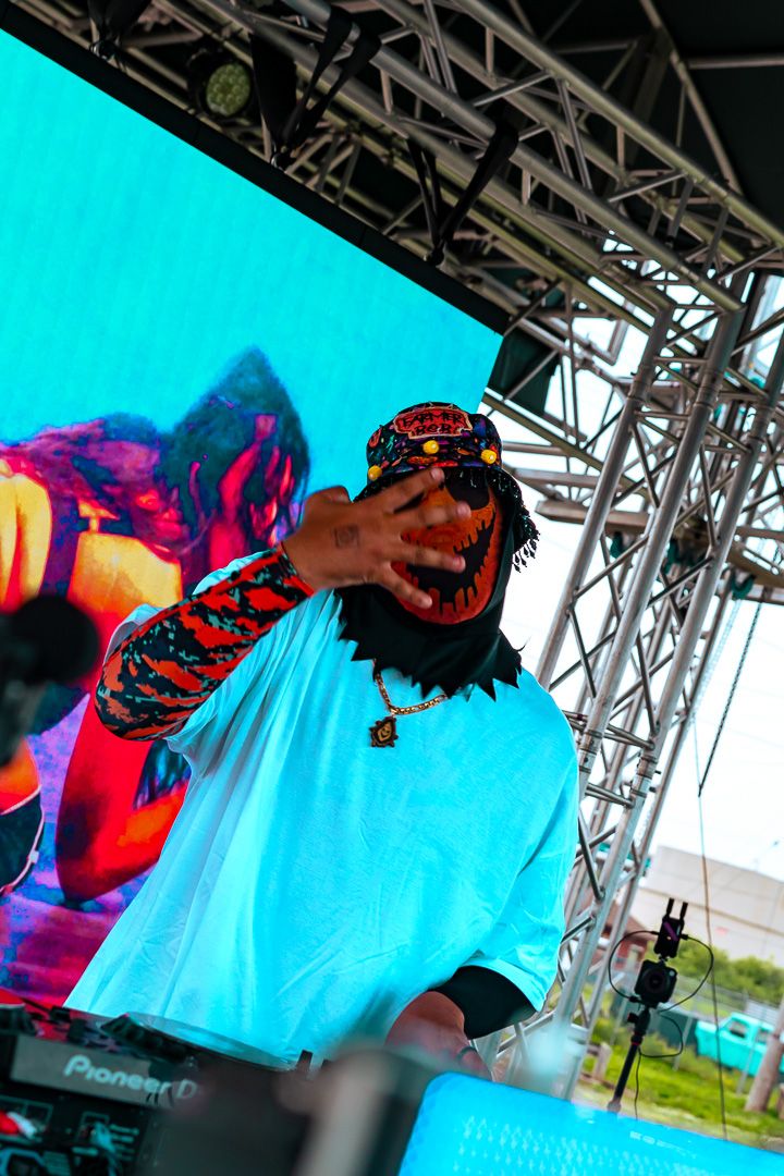 A DJ performer in colorful attire gestures on stage with Pioneer DJ equipment and a vibrant LED screen backdrop.