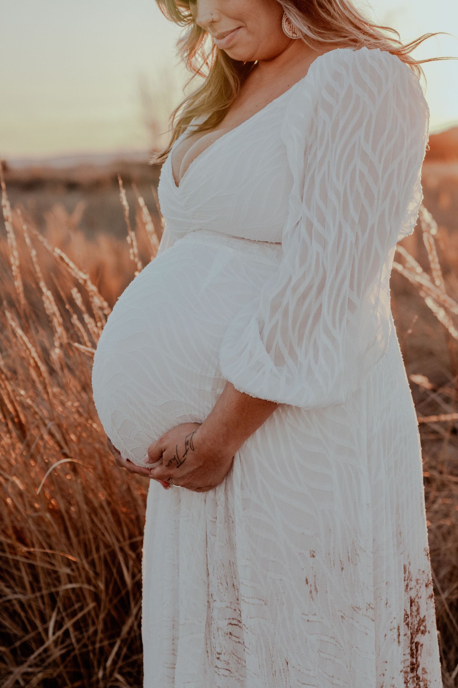 Pregnant woman in white lace maxi dress gently holding her baby bump in a golden sunlit field at sunset.