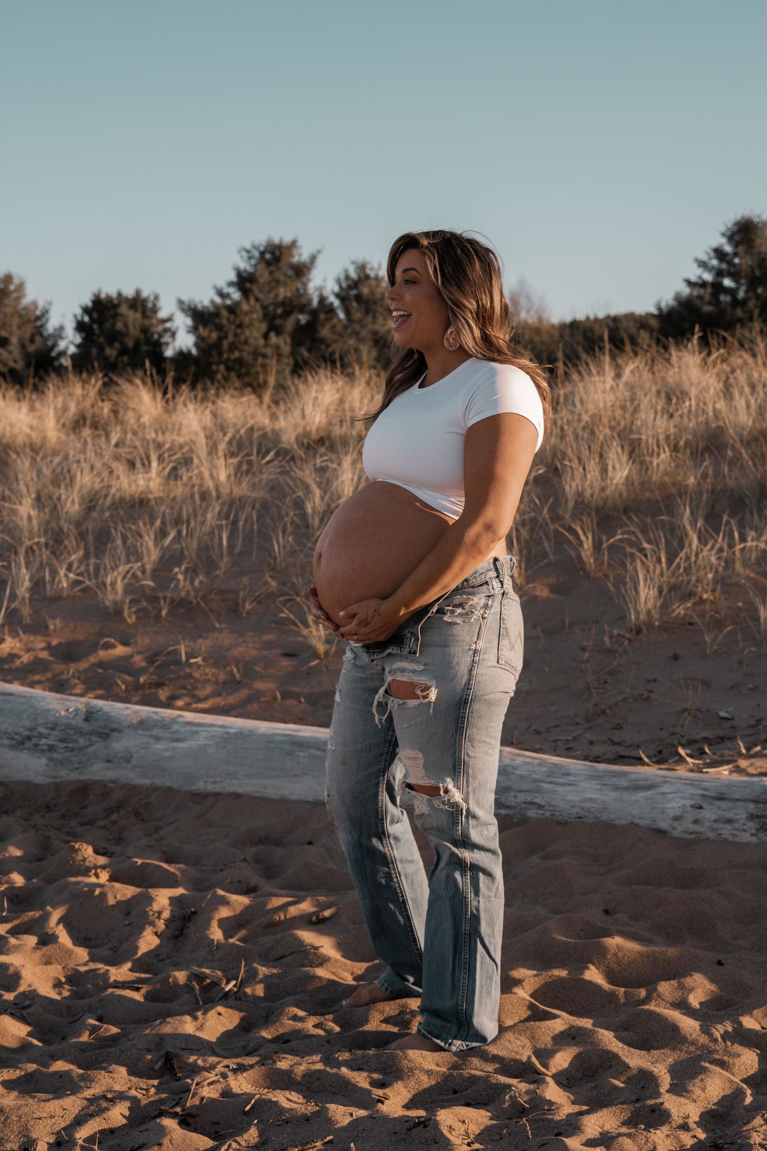 Smiling pregnant woman in white crop top and ripped jeans cradling her baby bump on a sandy beach at golden hour.