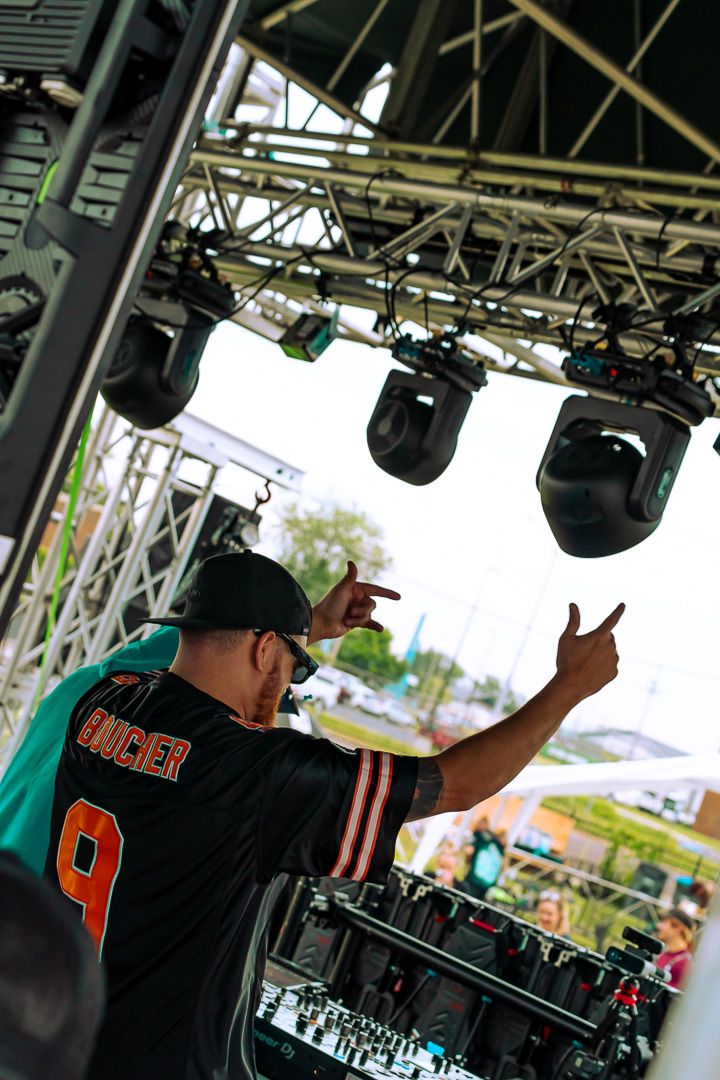 DJ performing on outdoor stage wearing jersey number 9, pointing fingers up under stage lighting rigs.