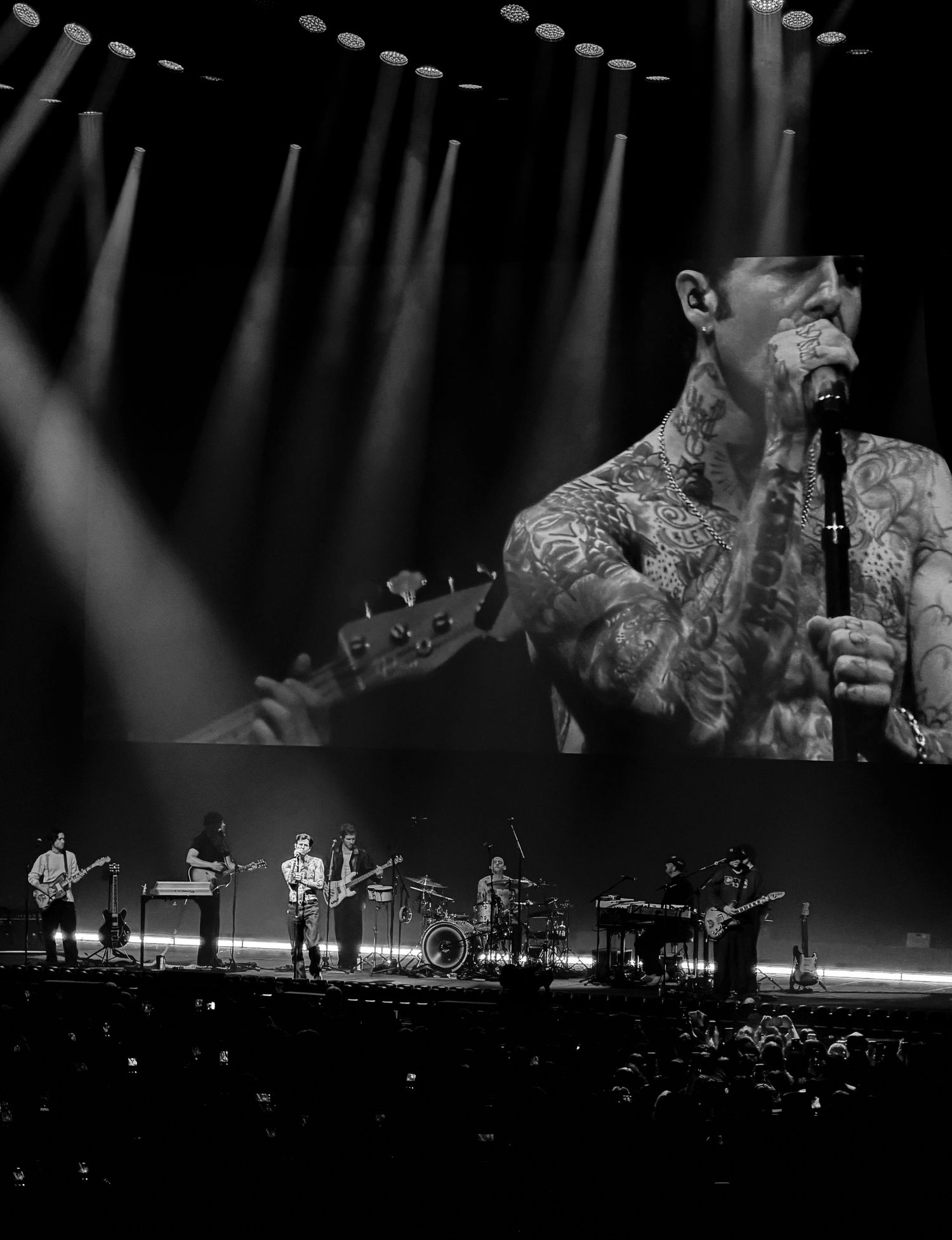 Black and white concert photo of a tattooed singer on screen above a live band performing on a dramatic lit stage.
