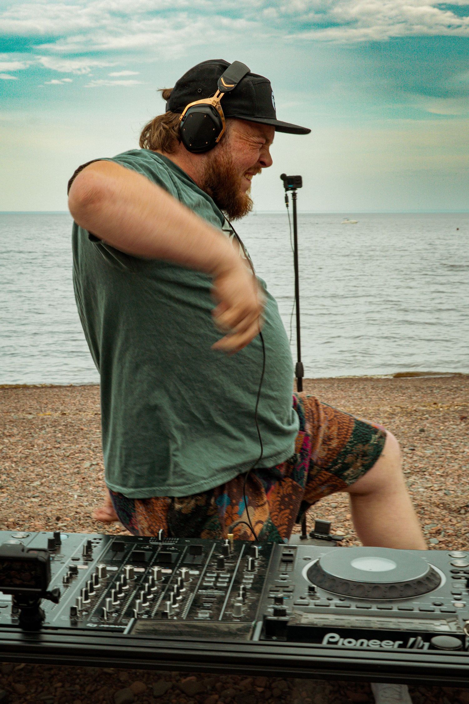 Bearded DJ with headphones dancing behind Pioneer DJ decks on a pebble beach with ocean backdrop.