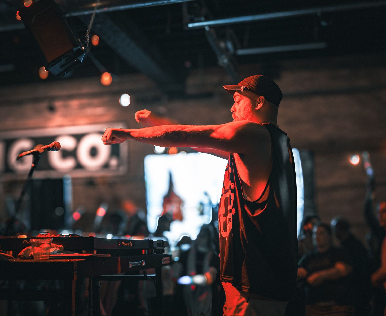 A DJ in a tank top and cap points at the crowd while performing at a dimly lit venue with warm red lighting.