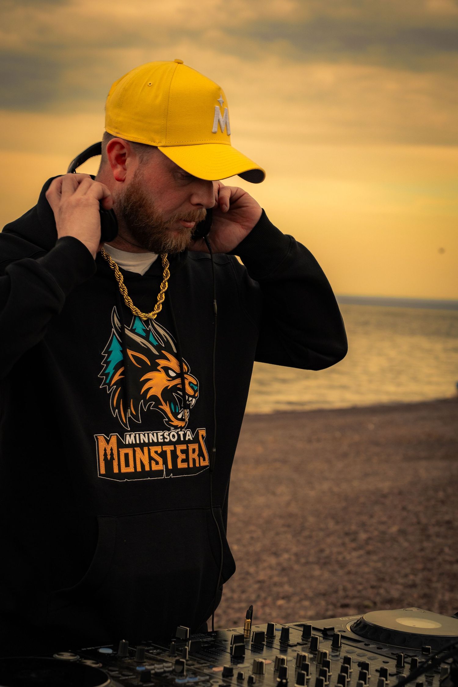 DJ wearing Minnesota Monsters hoodie and yellow cap mixing at outdoor beach setup during golden hour sunset.