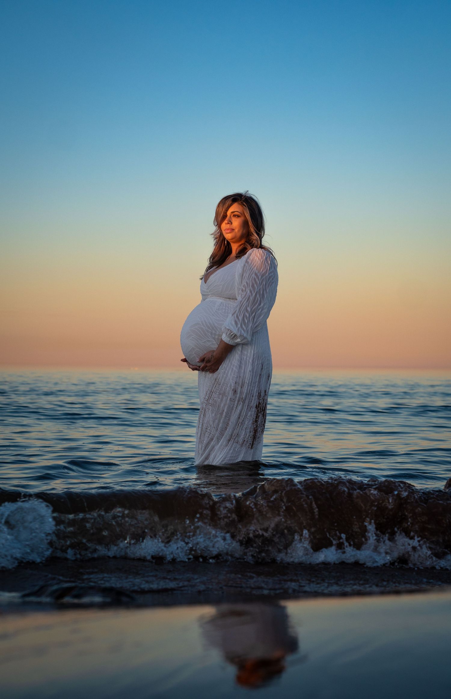 Pregnant woman in white lace dress standing in ocean waves at sunset, cradling her baby bump with reflection below.