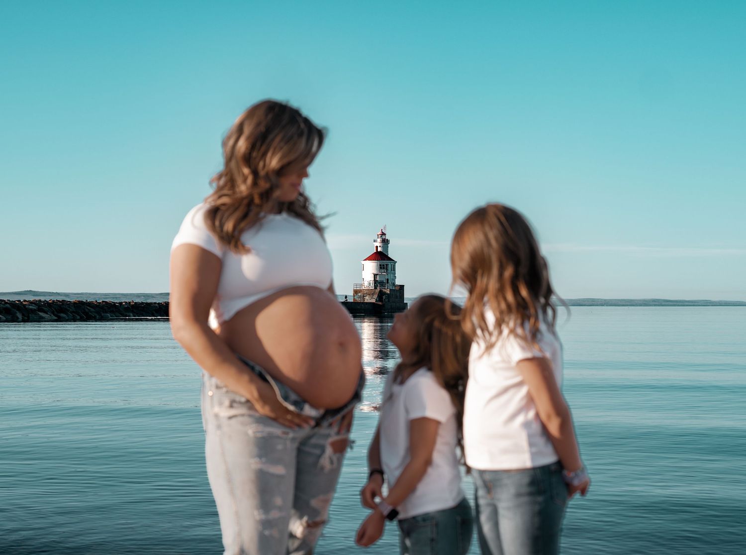 Pregnant woman and two children standing by the water near a lighthouse on a clear sunny day.