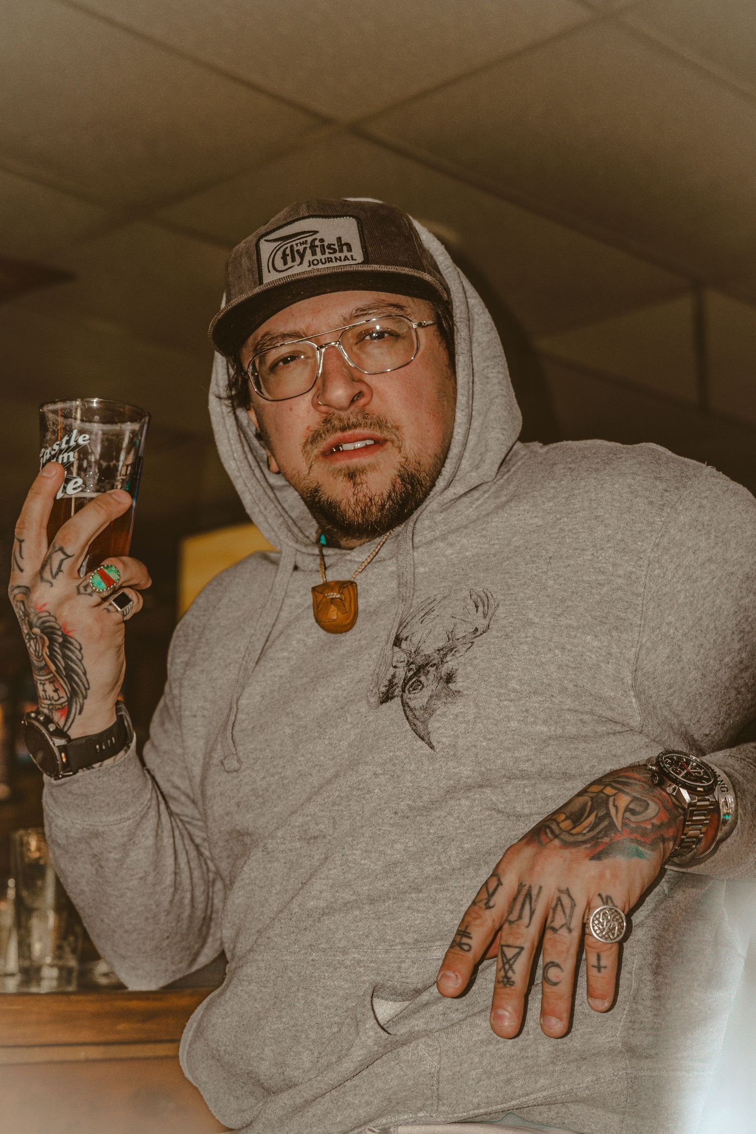 Tattooed man in gray hoodie and trucker hat holding a beer glass, wearing rings and a necklace indoors.