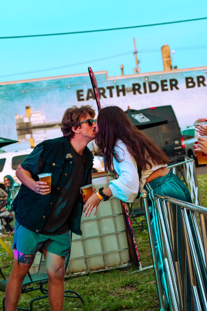 Couple kissing at Earth Rider Brewery outdoor festival, man holding beer, woman in teal pants, festive summer event.