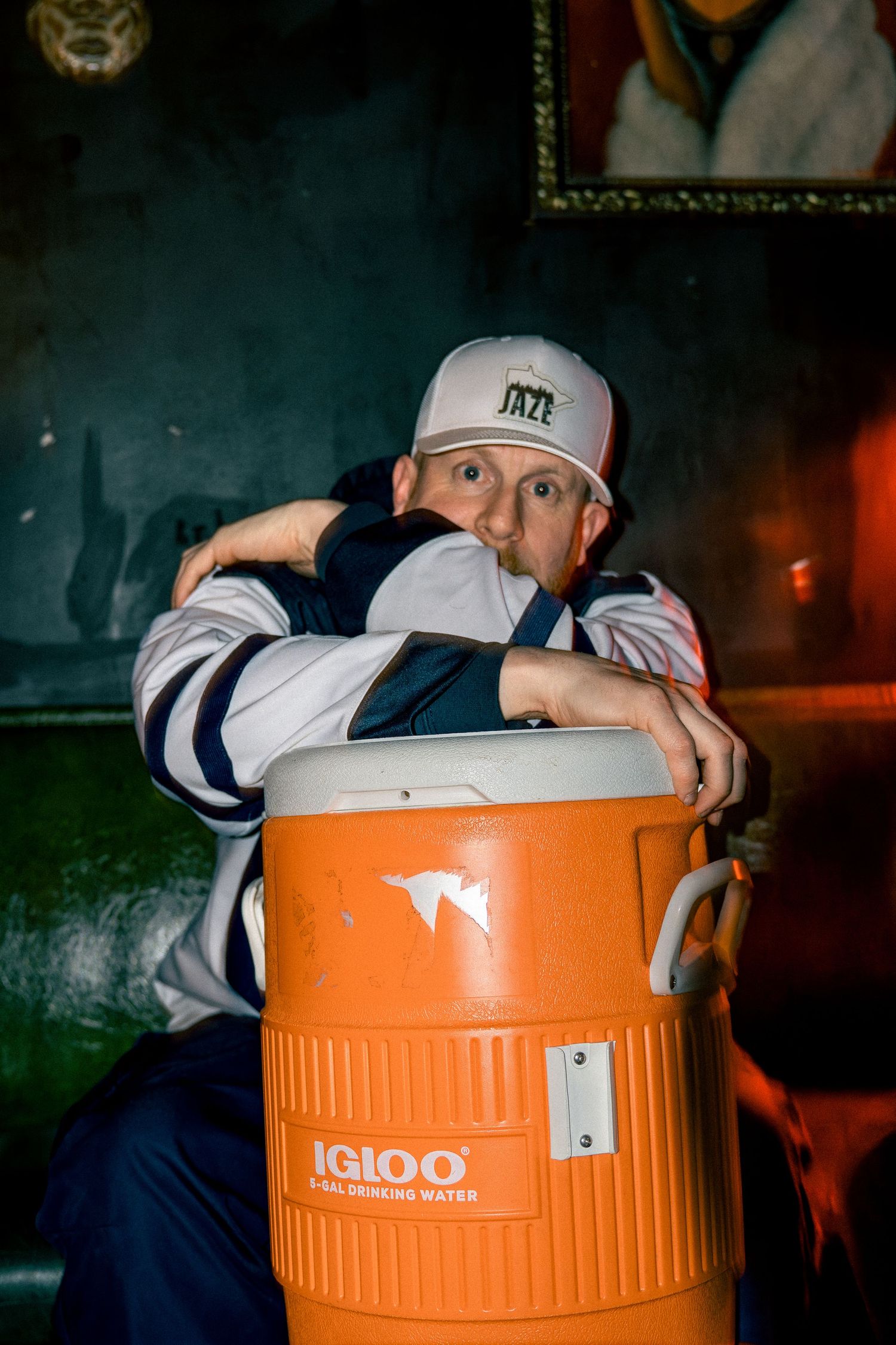 Man in a white Jazz cap leans on an orange Igloo water cooler in a dimly lit venue.