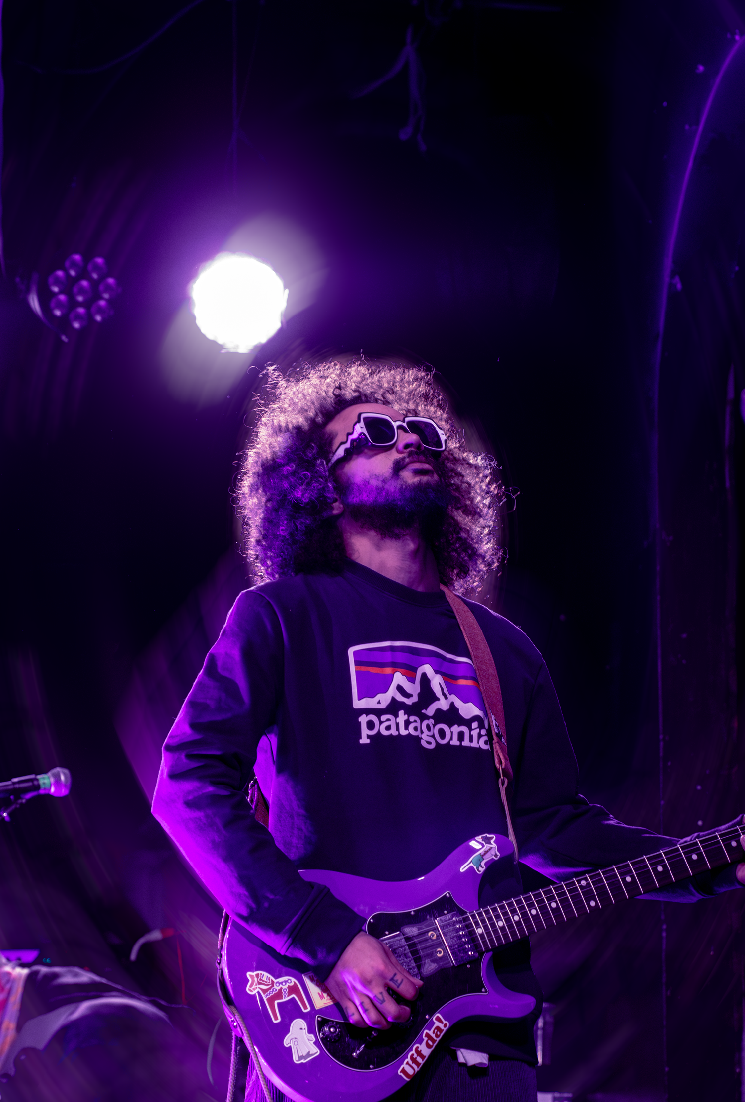 Curly-haired guitarist in Patagonia sweatshirt and sunglasses performs on purple-lit stage under bright spotlight.