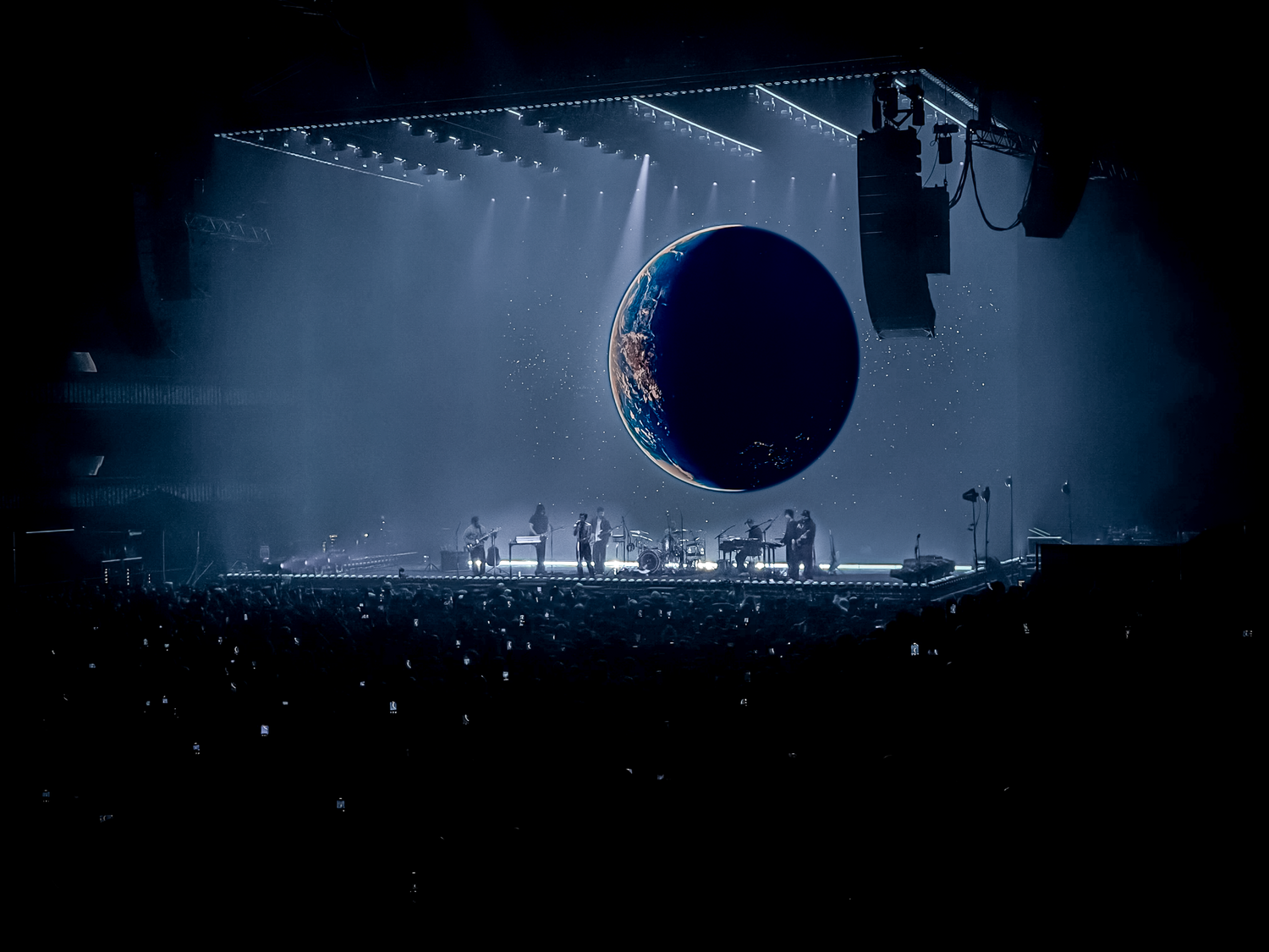 A massive glowing Earth sphere hovers above a dark concert stage with dramatic blue lighting and silhouetted performers.