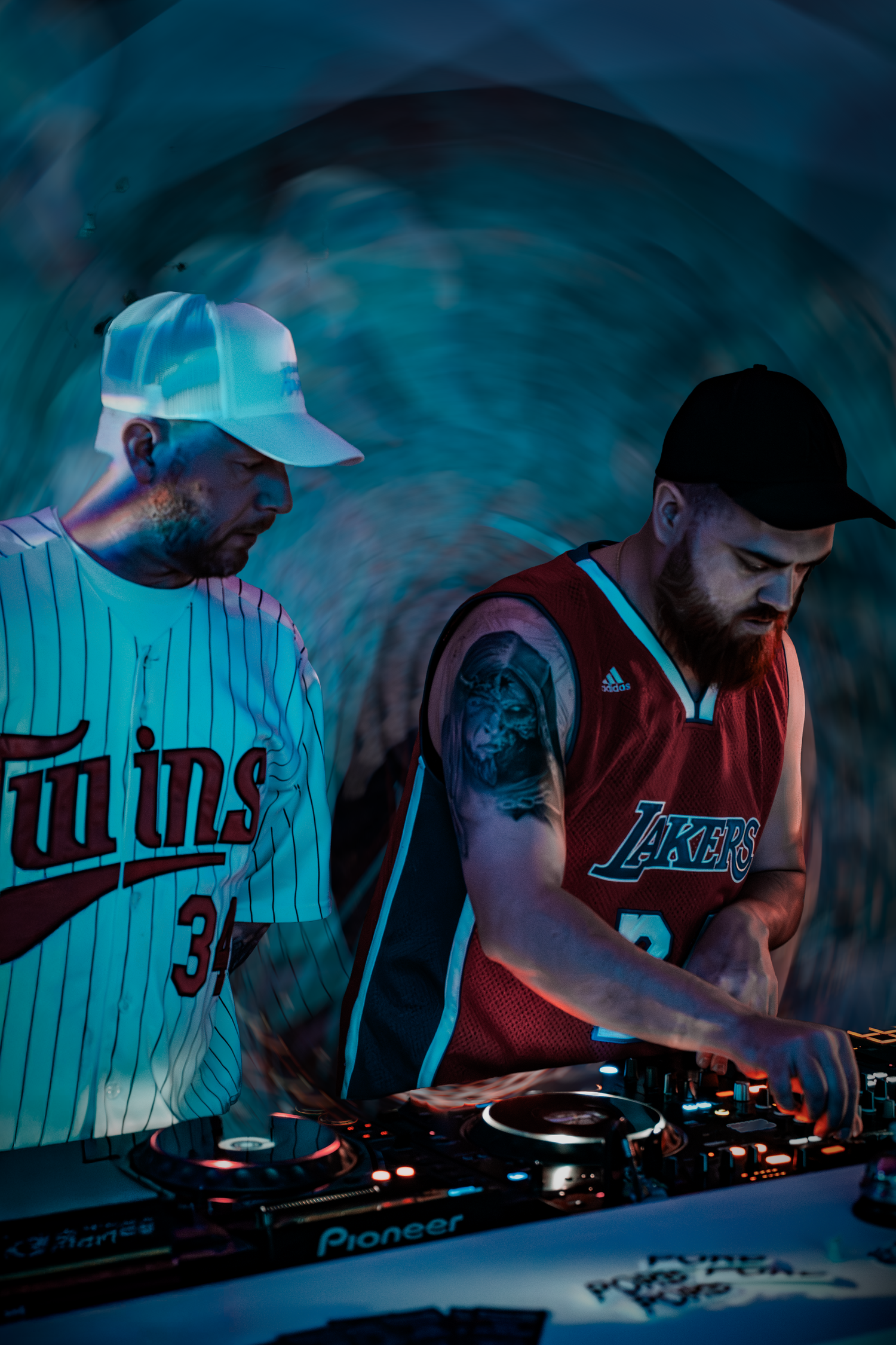 Two DJs in sports jerseys mixing on Pioneer decks under blue stage lighting at a music event.