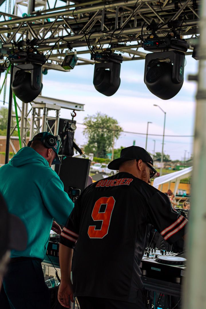 Two DJs perform on an outdoor stage under professional lighting rigs at a live music event.
