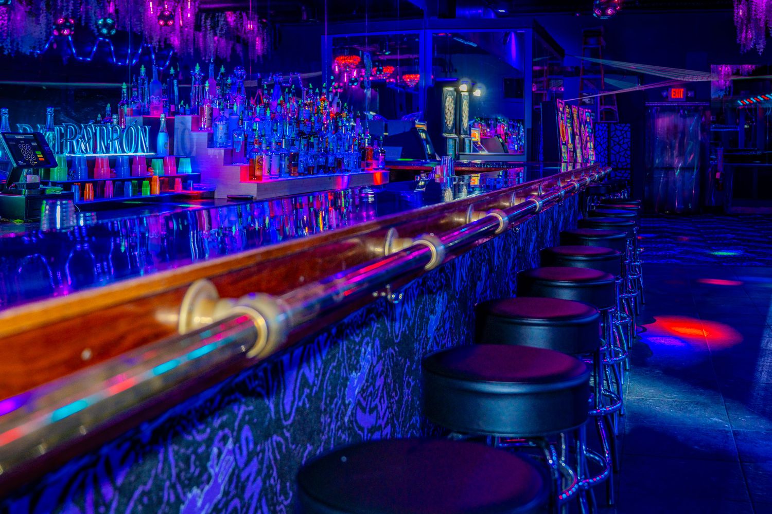 Neon-lit nightclub bar with blue lighting, illuminated liquor bottles, and black round bar stools along a glossy counter.