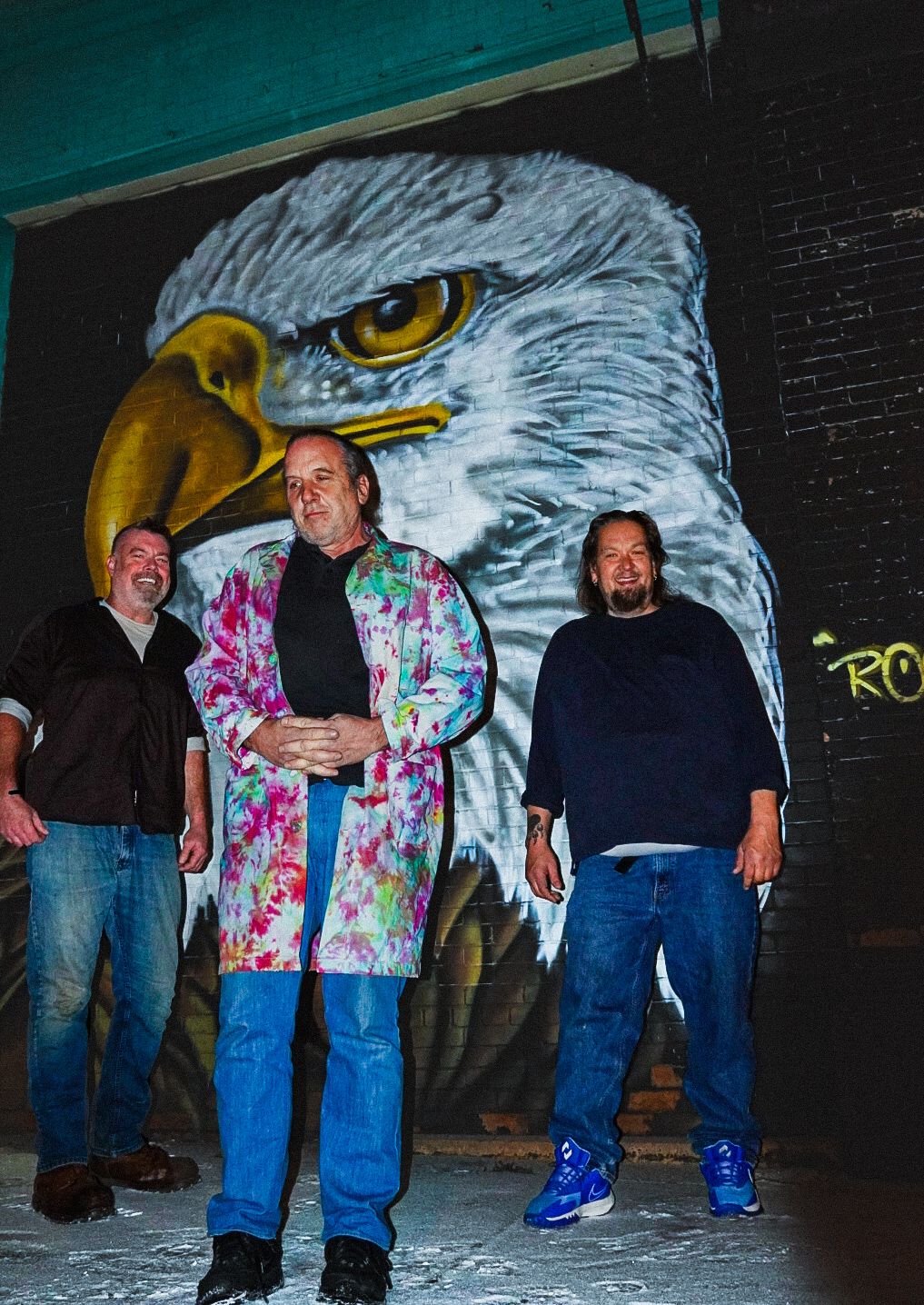 Three men pose in front of a large eagle mural on a brick wall, one wearing a colorful tie-dye jacket.