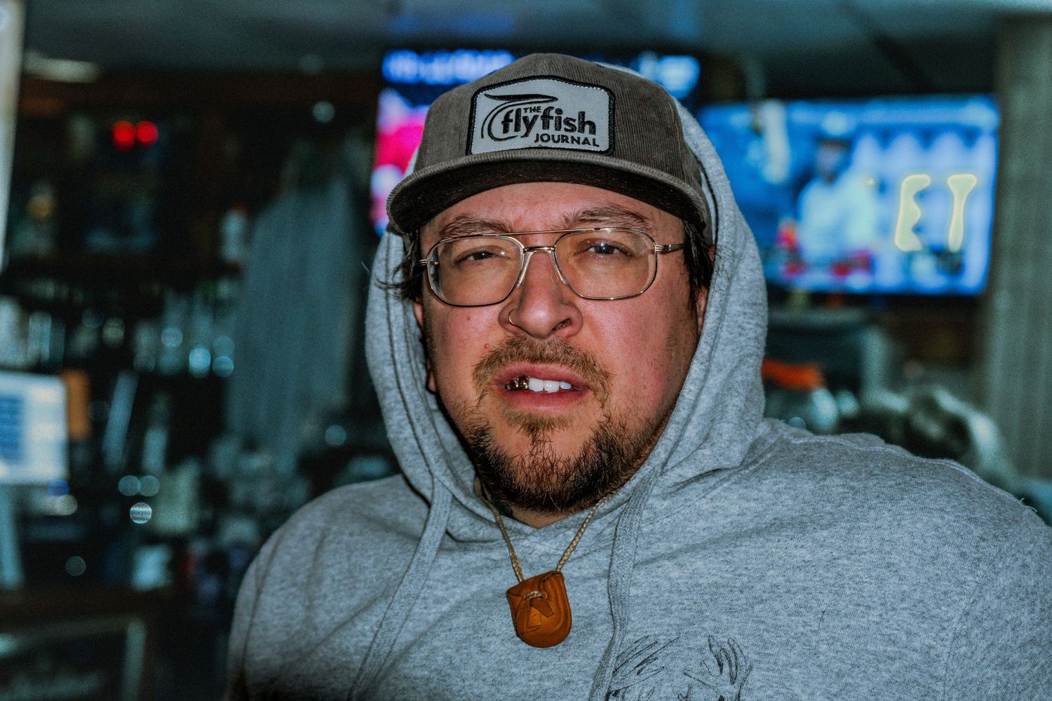 Man wearing gray hoodie and Flyfish trucker hat with glasses and beard poses indoors.