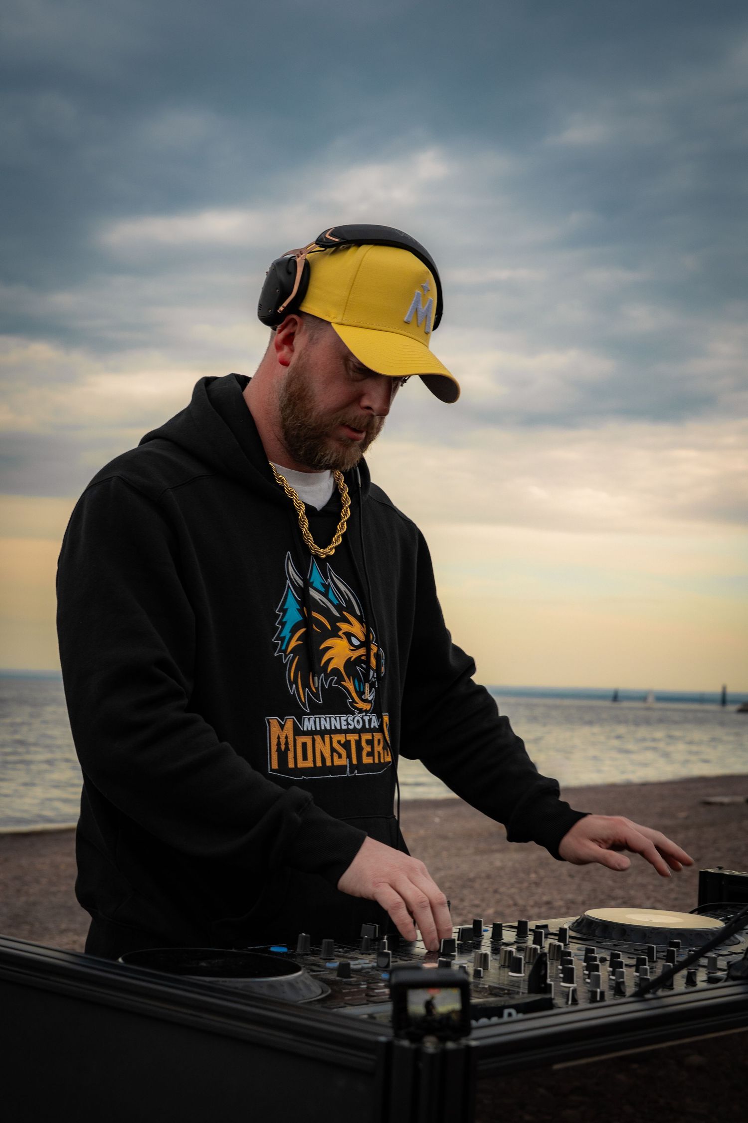 DJ in yellow cap and Minnesota Monsters hoodie mixing at outdoor turntables by the ocean at sunset.