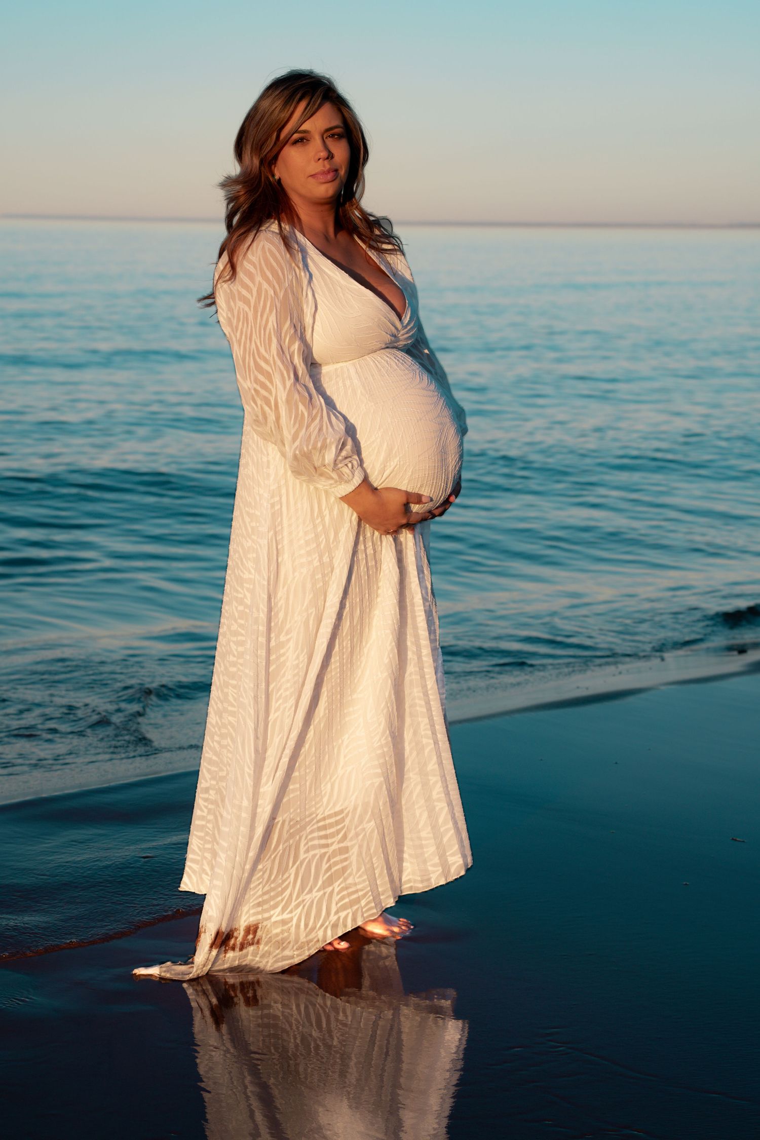 Pregnant woman in white dress standing barefoot on beach shore, cradling baby bump with ocean waves behind her.